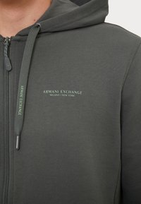 Haut à capuche gris zippé en tissu doux, doté d'une capuche, d'un logo contrastant vert "Armani Exchange Milano/New York" et d'un cordon de serrage texturé.