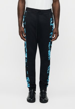 Man wearing black pants with blue marble-patterned side panels, black sneakers, and a white shirt, standing against a plain background.