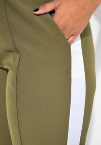 Olivově zelené legíny vyrobené z hladké tkaniny, které mají bílý boční pruh a boční kapsu. Design je přiléhavý a má elegantní siluetu.