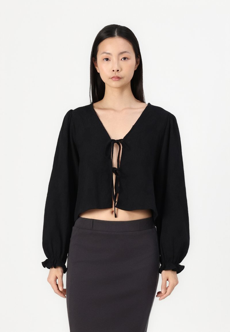 Blusa nera corta con scollatura a V profonda, maniche lunghe a palloncino e chiusura con lacci sul davanti. Realizzata in tessuto leggero con una consistenza morbida.