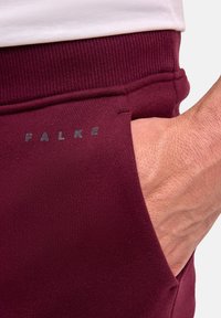 Bordeauxfarbene Sweatpants mit geripptem Bund, ausgestattet mit einerseitlichen Tasche und strukturiertem Stoff. Der Markenname "FALKE" ist in Grau gedruckt.