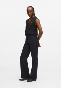 Must värviga ilma varrukateta ülikond V-kaelusega, koos kohandatud mustade pükste ja elegantselt teravate põranda-kingadega. Riietuse detailide seas on peen logo.