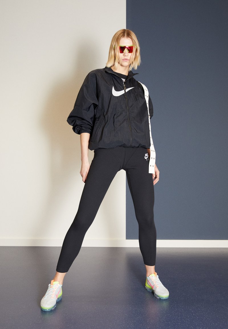 Črna lahka jakna z Nike logotipom, v kombinaciji s črnimi legicami in večbarvnimi športnimi čevlji. Model nosi rdeče sončne očala.