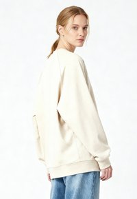 Felpa oversize beige chiaro con scollo rotondo, spalle scese, polsini e orlo a costine. Indossata con jeans di denim blu.