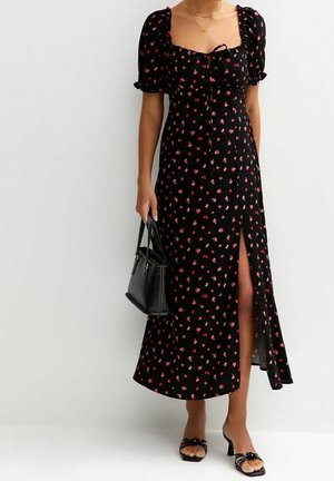 Vestido maxi negro con estampado floral, mangas cortas abullonadas, escote cuadrado y abertura lateral. Combinado con un pequeño bolso negro y sandalias transparentes de tacón.