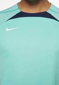Camiseta deportiva Nike en turquesa con acentos en verde texturizado en los hombros y una yugo en azul marino. Presenta un logo de swoosh blanco en el pecho.