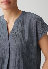 Blusa grigia con texture, scollo a V, maniche corte e sottili pieghe. Il materiale appare leggero e leggermente stropicciato.