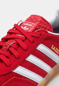 Červené semišové tenisky s třemi bílými pruhy, gumovou podrážkou a texturovaným červeným jazykem s logem. Zlaté "GAZELLE" po straně.