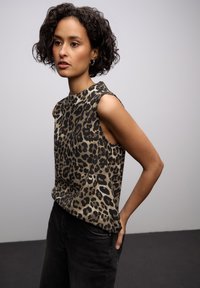 Top bez rękawów w panterkę w beżowym i czarnym kolorze; z wysoką szyją i luźnym krojem, zestawiony z ciemnymi dżinsami.
