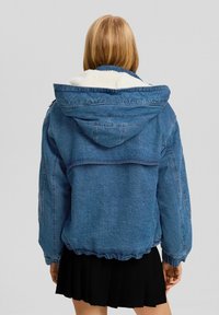 Veste en denim bleue avec capuche, dotée d'une doublure blanche duveteuse, de poignets élastiques et d'un ourlet resserré. La vue de dos montre un design simple et des poches.
