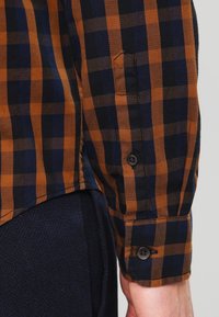 Chemise à carreaux marron et bleu à manches longues, avec un détail de bouton au poignet et un tissu texturé. La coupe semble décontractée.