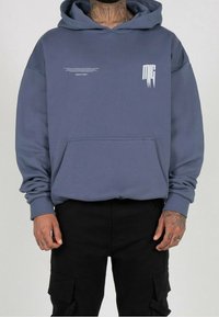 Mies, jolla on päällään sininen huppari, jossa on valkoista tekstiä rinnassa, ja mustat cargo-housut, seisoo kädet rentoina sivuilla.