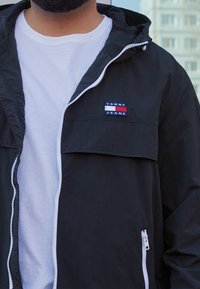 Czarna kurtka z kapturem i zamkiem błyskawicznym z przodu, z białymi akcentami na zamku oraz kolorową naszywką z napisem "TOMMY JEANS". Noszona na białej koszulce.