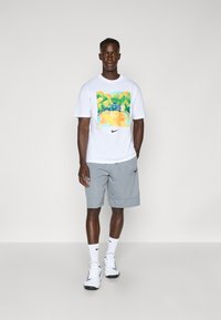 Hvid t-shirt med farverig grafikdesign featuring grønt, parret med grå shorts. Båret med hvide sneakers og sokker, minimal branding synlig.