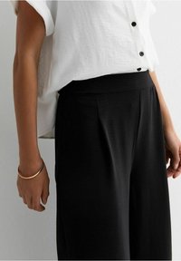 Pantalons larges noirs en matériau lisse avec un devant plissé, associés à un haut blanc à manches courtes et à boutons.