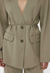 Kakija žakete ar dziļu V veida izgriezumu, platiem piedurknēm, divām priekšējām kabatām un jostu viduklī. Priekšpusē divas pogas.