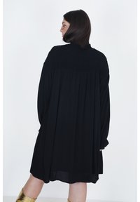 Zwarte, oversized jurk met een smock-bodice, lange mouwen en een vloeiende silhouet. Stof lijkt licht en zacht. Gouden metallic laarzen worden gedragen.