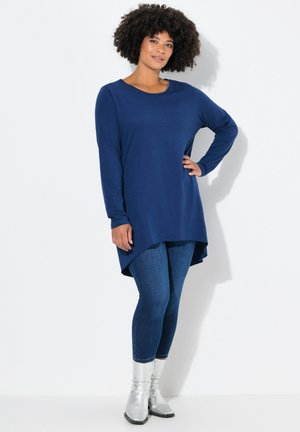 Marineblauwe tuniek met lange mouwen en een asymmetrische zoom, gecombineerd met donkere denim legging en zilveren enkellaarsjes op een neutrale achtergrond.