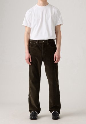 Osoba ubrana w gładki biały t-shirt wpuszczony w ciemnobrązowe sztruksowe spodnie i czarne skórzane buty, stojąca na jasnym neutralnym tle.