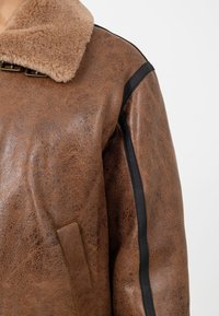 Braune Kunstlederjacke mit einem plüschigen beigen Kragen, mit schwarzen seitlichen Akzenten und einem strukturierten Design. Enthält ein seitliches Taschendetail.