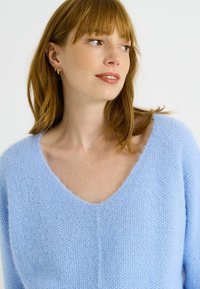 Pull en tricot bleu clair avec un décolleté en V profond, texture douce et coupe loose. Présente une apparence subtile et duveteuse avec des épaules légèrement tombantes.