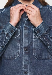 Camisa de mezclilla en azul oscuro con cuello clásico, cierre frontal de botones y dos bolsillos en el pecho. La textura está ligeramente desvanecida con costuras visibles.