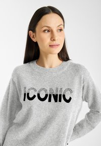 Grijs sweater gemaakt van zacht materiaal met het woord "ICONIC" in vette zwarte letters, versierd met een strassversiering op een deel van de tekst.