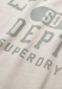 Primo piano di un tessuto bianco con testo grigio sbiadito che recita "SD DEPT SUPERDRY" e una grafica parziale di una chiave.