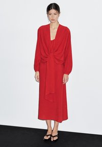 Rode jurk van gemiddelde lengte met lange mouwen, gathered tie-detail in de taille en een soepele textuur. Draag het met eenvoudige zwarte sandaal.