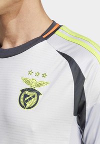 Camiseta deportiva blanca con detalles en gris, que presenta franjas amarillas y naranjas en los hombros, y un destacado emblema amarillo del club con un águila.