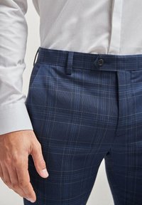 Homme portant un pantalon ajusté à carreaux bleu marine et une chemise blanche à manches longues, montrant les détails des boutons et des passants de ceinture près de la taille.