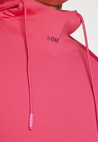 Różowy sweter z wysokim kołnierzem i sznurkiem, z logiem "BOSS" w ciemnym druku. Gładka tekstura i nowoczesne detale designu.