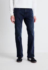 Jeans de mezclilla azul oscuro con corte recto, diseño de cinco bolsillos y un sutil desvanecimiento en el frente. Llevados con zapatillas negras con rayas blancas.