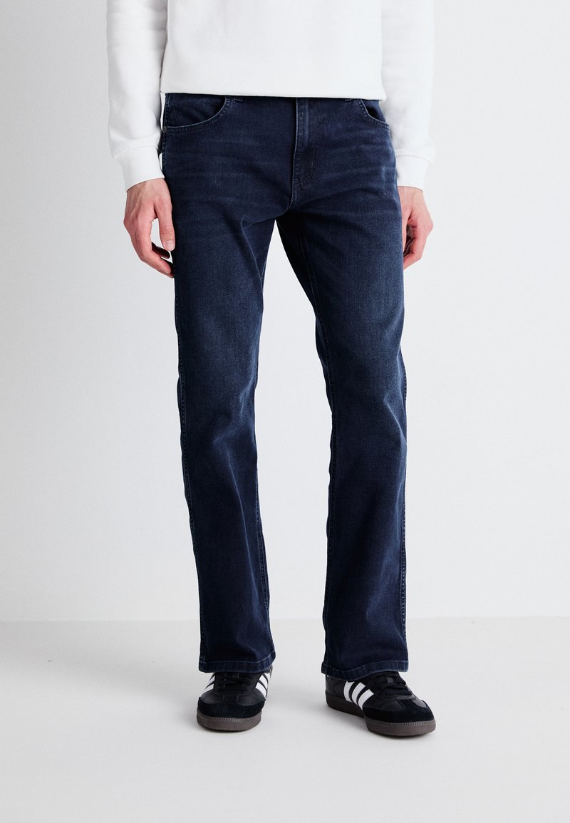 Jeans de mezclilla azul oscuro con corte recto, diseño de cinco bolsillos y un sutil desvanecimiento en el frente. Llevados con zapatillas negras con rayas blancas.