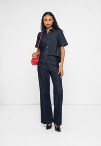 Mulher vestindo uma camisa de manga curta em denim escuro e calças largas, segurando uma pequena bolsa vermelha a tiracolo, em pé contra um fundo branco.