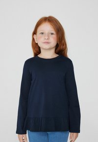 Granatowa, długosleeve'owa koszulka z okrągłym dekoltem i falbankowym dołem. Wykonana z miękkiego materiału i o luźnym kroju. Połączona z jasnoniebieskimi dżinsami.