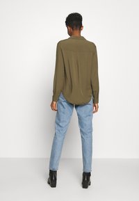 Oliivinvihreä pitkähihainen pusero pehmeästä kankaasta, jossa on takana laskos ja pyöristetty helma, yhdistetty vaaleansinisiin suoralahkeisiin farkkuihin ja mustiin saappaisiin.