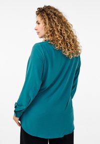 Türkisfarbenes Langarmshirt mit abgerundetem Saum und hochgerollten Ärmeln. Glatte Stoffqualität mit lockerer Passform, keine sichtbaren Muster oder Hardware-Details.