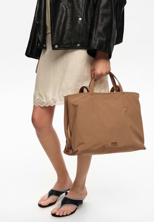 Mujer con chaqueta de cuero negra y vestido crema con ribete de encaje, llevando un gran bolso tote color marrón claro, y sandalias negras abiertas en los dedos.