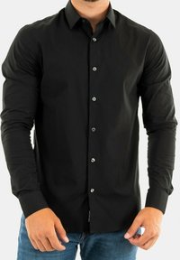 Mężczyzna w dopasowanej czarnej koszuli z guzikami i długimi rękawami, w niebieskich dżinsach, stojący na jednolitym tle.