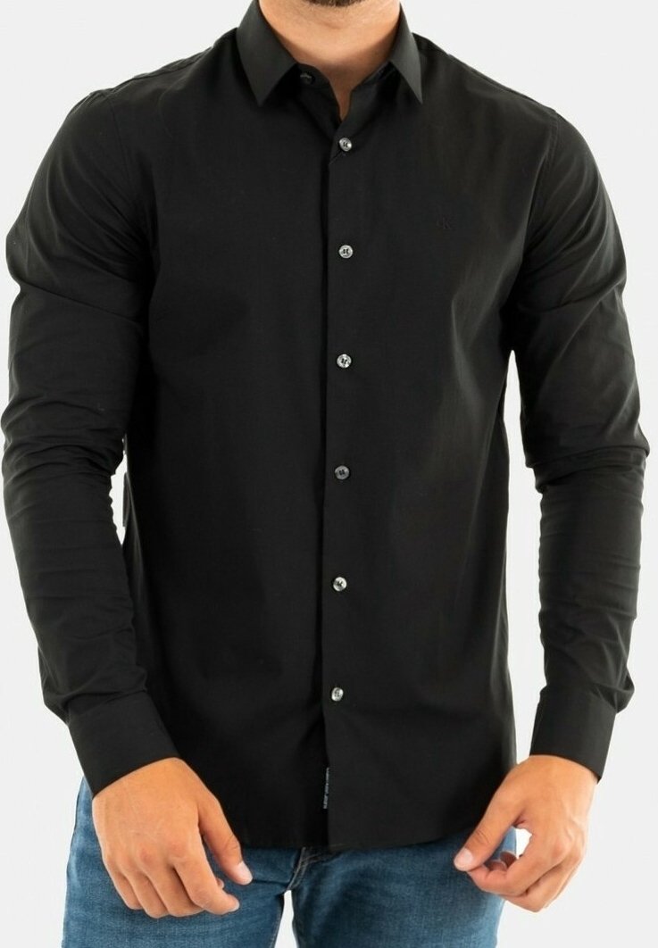Mężczyzna w dopasowanej czarnej koszuli z guzikami i długimi rękawami, w niebieskich dżinsach, stojący na jednolitym tle.