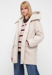 Abrigo de lana beige claro con un acabado texturizado, capucha oversize y cierre de botones a presión. Combinado con un suéter a rayas y jeans de mezclilla.