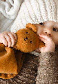 Ursuleț de pluș moale, maro, ținut de un copil care poartă un pulover texturat, de culoare crem, și o căciulă tricotată. Detalii apropiate vizibile.