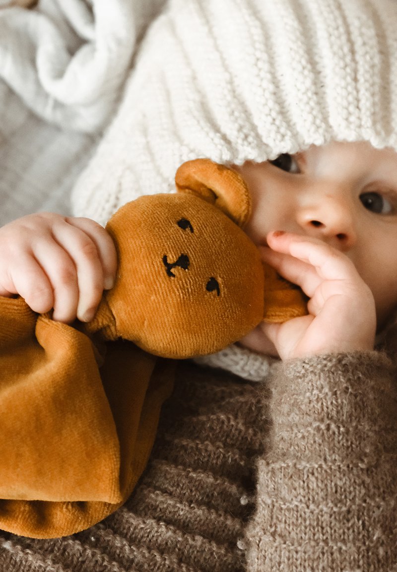 Ursuleț de pluș moale, maro, ținut de un copil care poartă un pulover texturat, de culoare crem, și o căciulă tricotată. Detalii apropiate vizibile.