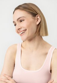 Jeune femme aux cheveux blonds courts souriante, portant un débardeur rose clair, bras croisés, sur un fond blanc uni.