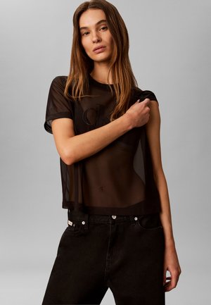 Žena s dlhými hnedými vlasmi, ktorá má na sebe priesvitné čierne tričko Calvin Klein a čierne nohavice, stojaca pred svetlosivým pozadím.