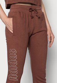 Brūnas kokvilnas bikses ar elastīgu jostasvītru, regulējamu auklu, kabatām un spīdīgu sudraba Adidas logotipa detaļu kreisajā kājā.