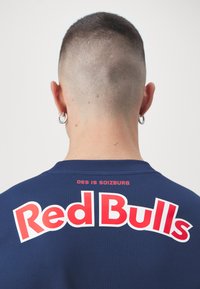 Maglietta sportiva blu navy con testo rosso "Red Bulls", maniche corte e texture a rete. "DES IS SOIZBURG" stampato sopra il logo principale.