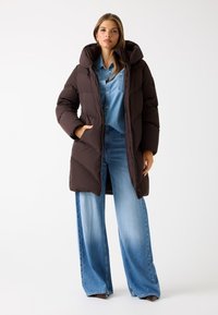 Bruine fluffy jas met een grote capuchon, met een gequilt ontwerp. Gdragen over een denim shirt en wijde blauwe jeans.