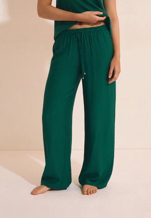 Osoba nosząca luźne spodnie w ciemnozielonym kolorze z elastycznym pasem i sznurkiem, boso na jasnej podłodze.